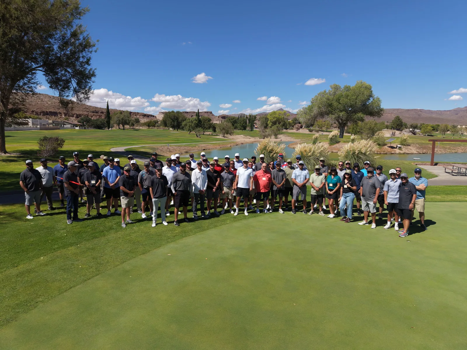 A large group poses on a golf green beside a pond, with trees and desert hills in the background.